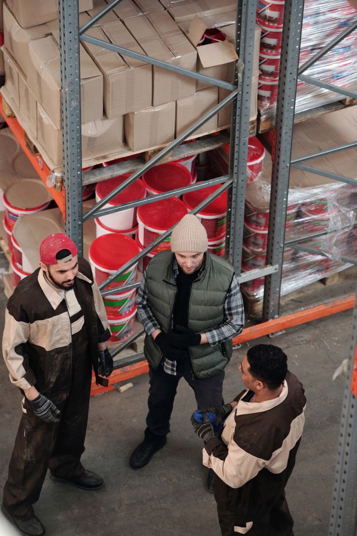 Group of warehouse workers discussing logistics in an industrial storage area with shelves and packaging.