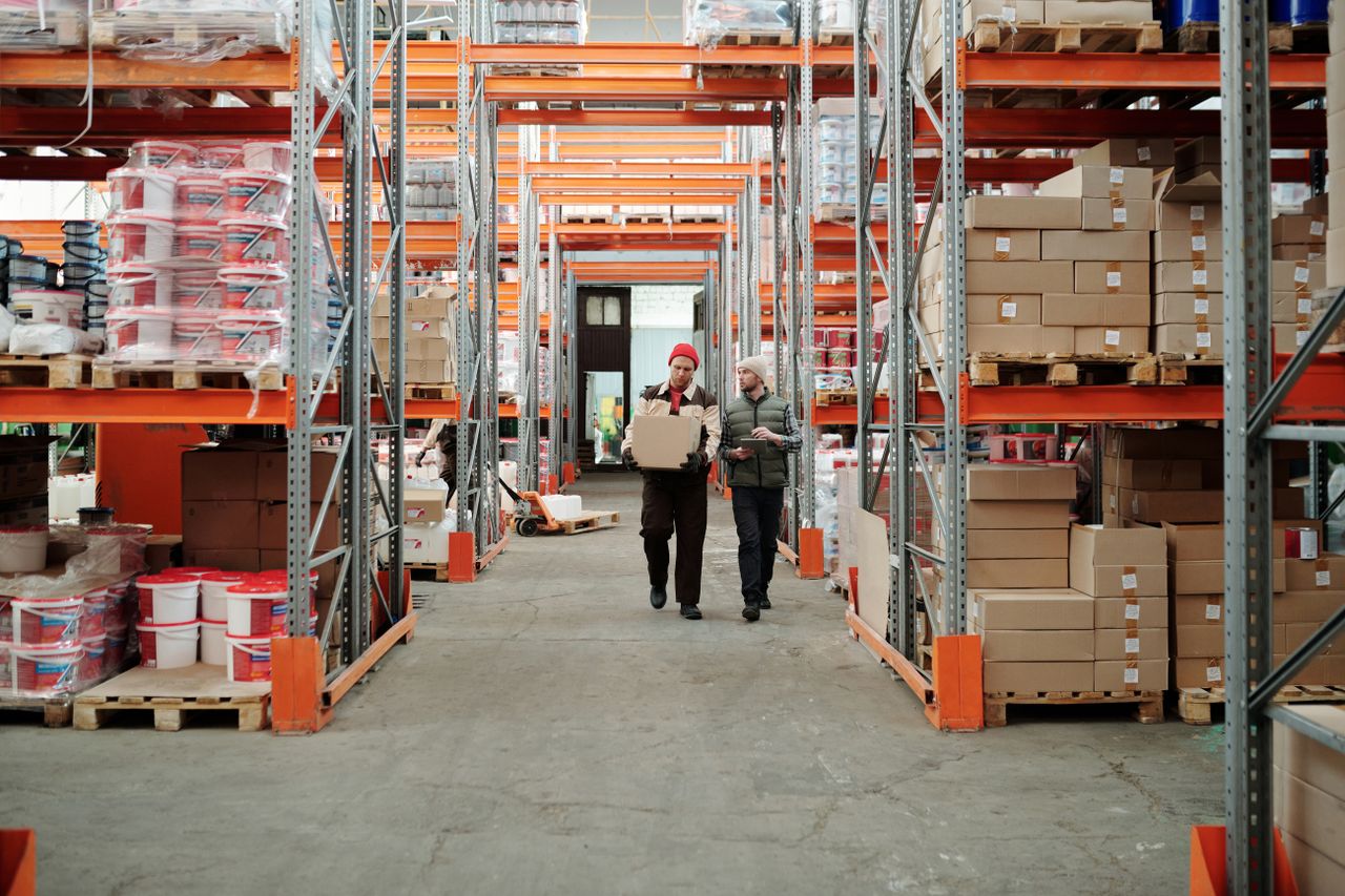 our-experience Two workers handle a package in a spacious warehouse surrounded by shelves stocked with boxes and products.